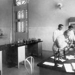 Oswaldo Cruz examina microscópio em laboratório de Manguinhos, observado por seu filho Bento Oswaldo Cruz e por Burle de Figueiredo
