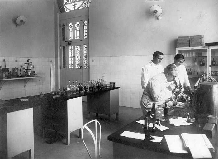 Oswaldo Cruz examina microscópio em laboratório de Manguinhos, observado por seu filho Bento Oswaldo Cruz e por Burle de Figueiredo